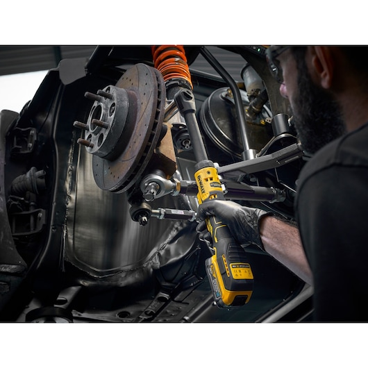 A person using a DEWALT DCF512EN cordless ratchet tool to tighten or loosen a bolt on the suspension system of a car. The tool is yellow and black, and the person is wearing black gloves.