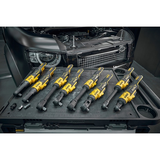 A set of Dewalt cordless ratchet tools displayed on a workstation, with a car engine and front grille visible in the background. Various tool attachments and sockets are also placed on the workstation.