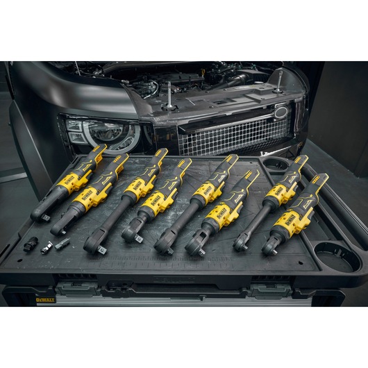Several yellow and black DEWALT cordless ratchet wrenches are arranged on a work surface in front of a partially opened vehicle hood, with other small tool accessories also visible.