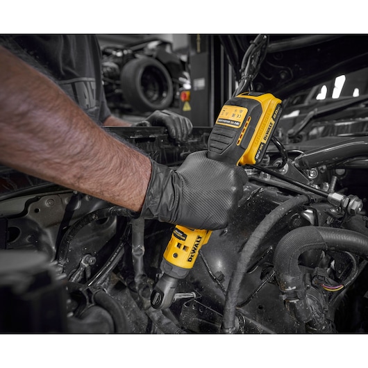 A person wearing black gloves uses a yellow DEWALT cordless ratchet tool to work on machinery or an engine in a workshop setting.