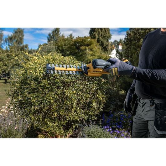 A person is using a DEWALT cordless hedge trimmer to shape a bush in a garden, surrounded by greenery and flowers.