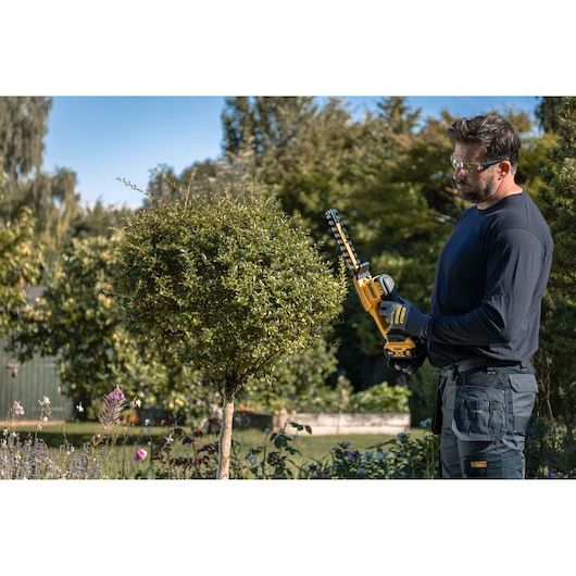 A person wearing gloves and outdoor clothing is using a yellow and black DEWALT cordless hedge trimmer to trim a small tree in a garden.