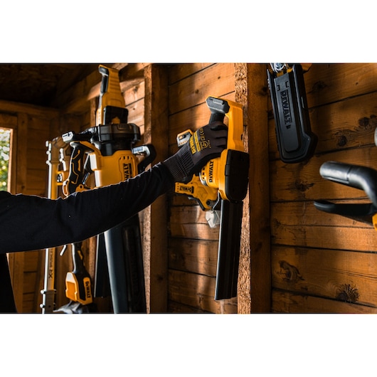 A person wearing gloves is hanging a DEWALT cordless hedge trimmer on a wooden wall in a tool shed. Other DEWALT tools are also mounted on the wall.