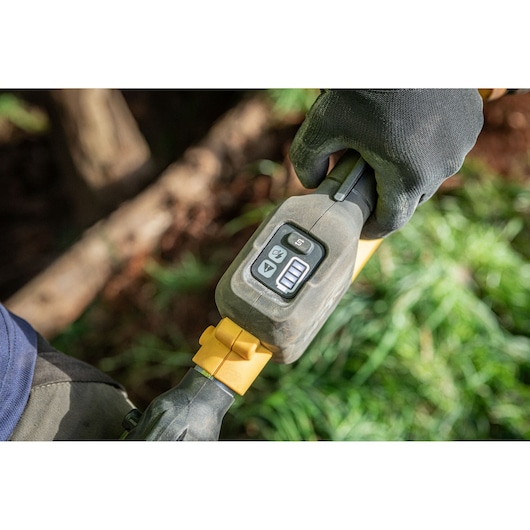 Close-up view of a gloved hand holding a DEWALT power tool with a control panel and digital display, outdoors near grass and soil.